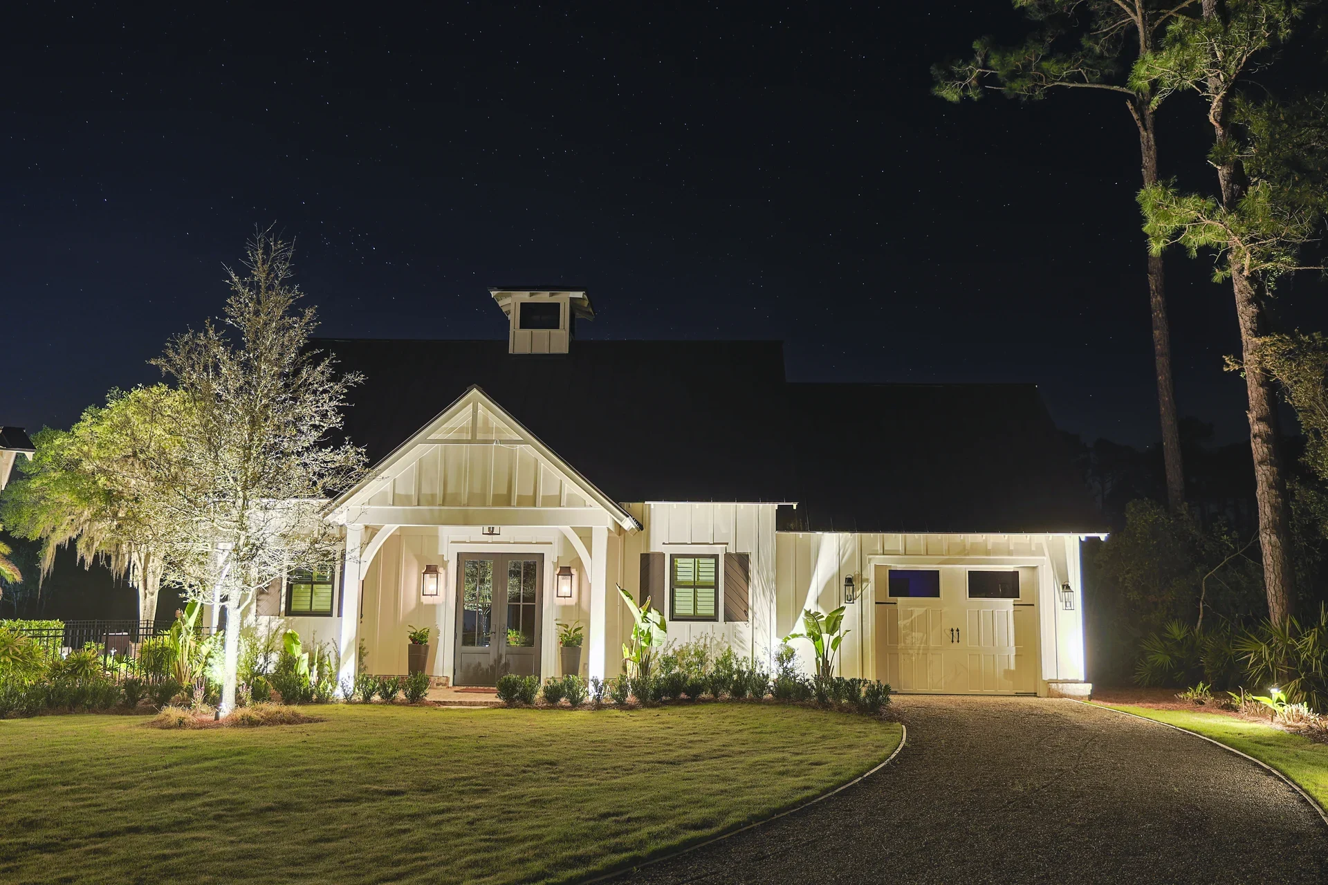Warm lighting illuminating a coastal home, trees, and a curved driveway at night